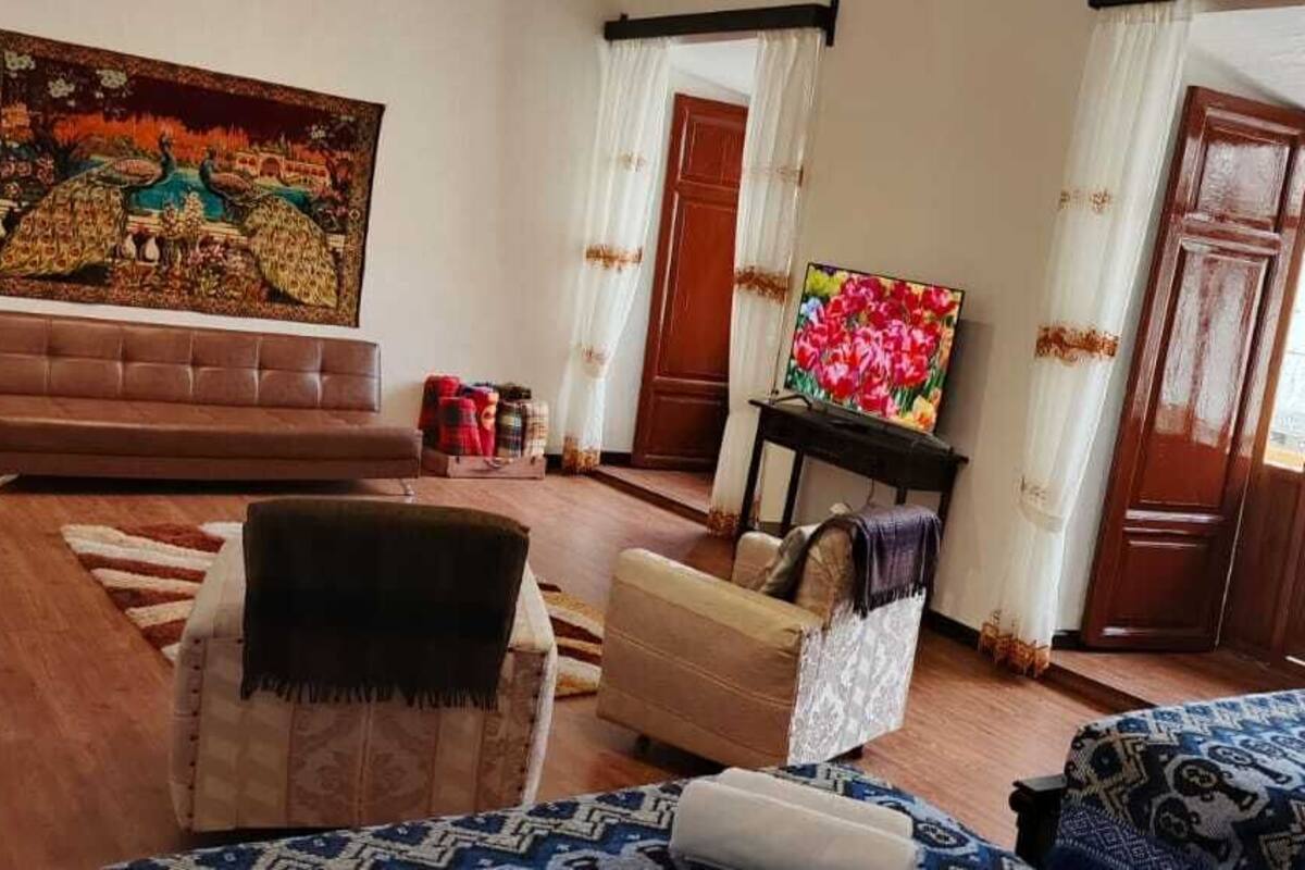 A living room with a sofa, armchairs, a coffee table, and a TV. There is a painting on the wall and a door leading to another room.