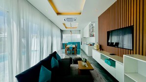 A modern living room with a sofa, coffee table, and TV. There is a dining area with a table and chairs in the background. - Kamala Garden View Villas by Aladdin (Kamala)
