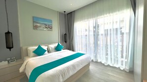 A modern hotel room with a large bed, white bedding, teal pillows, a painting on the wall, and a view through sheer curtains. - Kamala Garden View Villas by Aladdin (Kamala)