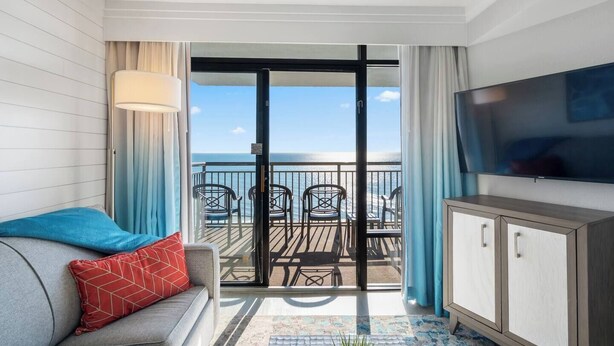En balkon med udsigt over havet, en sofa med en rød pude, et fjernsyn på væggen og et træskab.