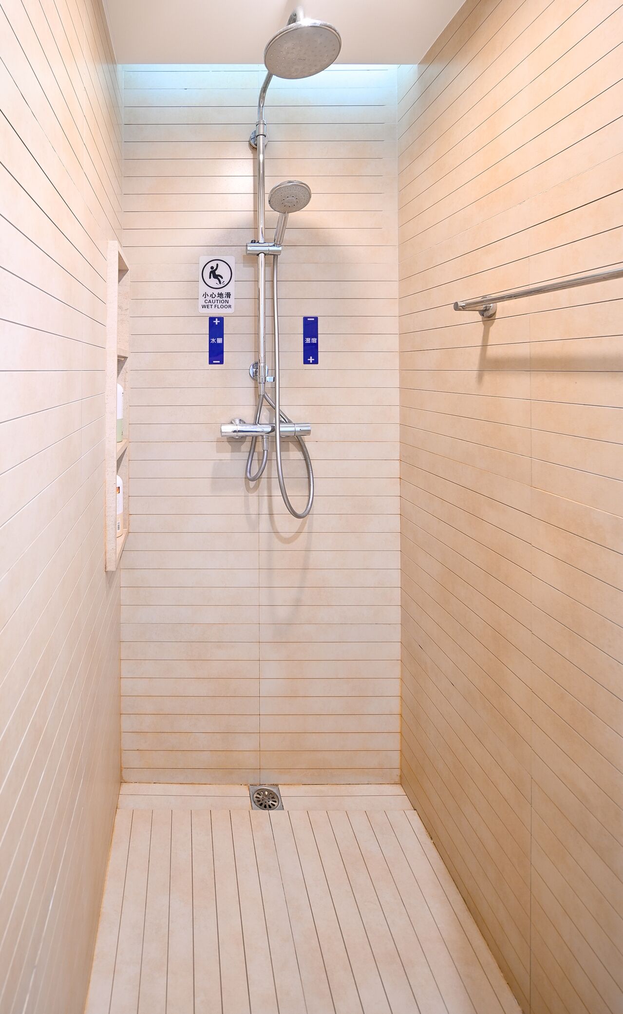 A small, tiled shower room with a handheld showerhead and a wall-mounted faucet. There are two signs indicating caution for wet floors and temperature control.