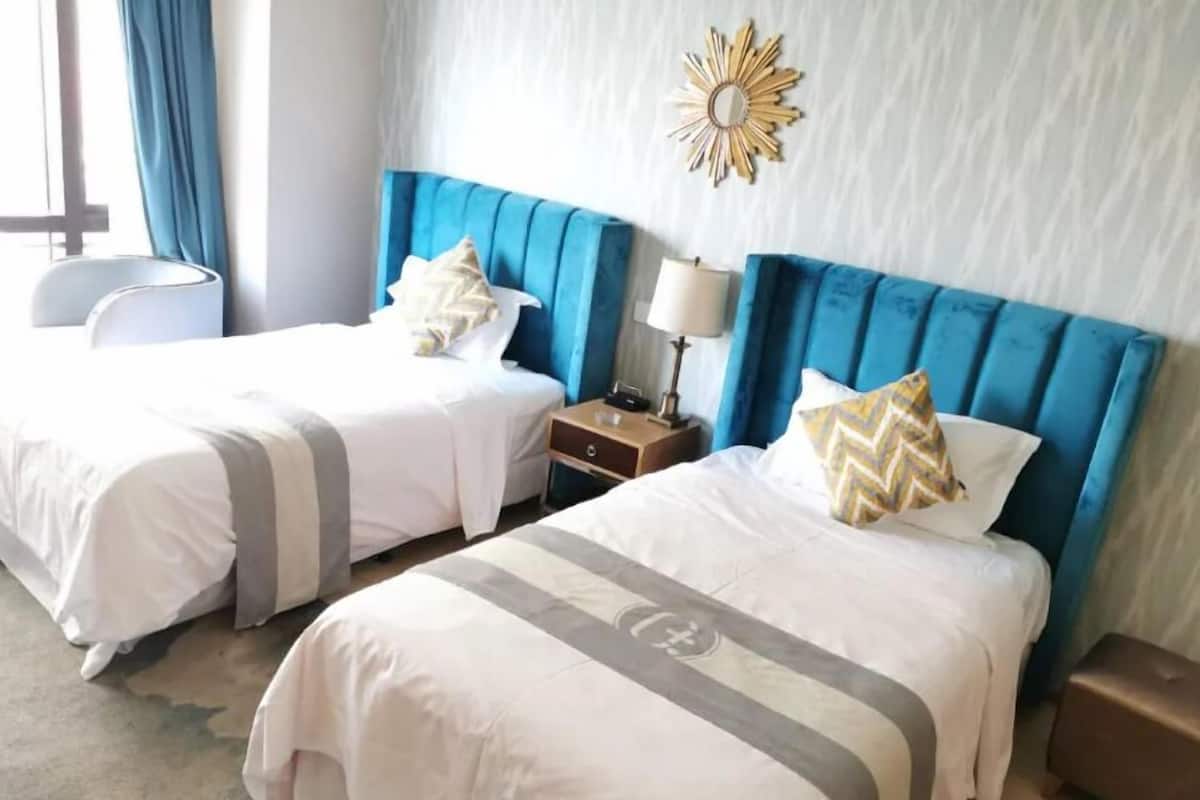 A hotel room with two beds, each with white bedding and blue headboards. There is a nightstand with a lamp between the beds, and a decorative mirror on the wall.