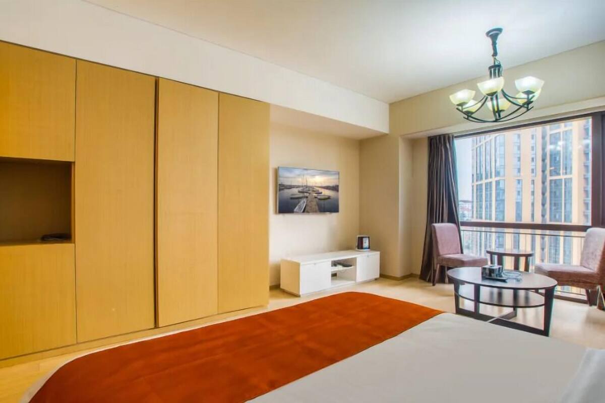 A hotel room with a large bed, a wooden wardrobe, a small white console table, a painting on the wall, a balcony with chairs, and a chandelier.