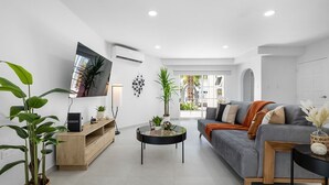 A modern living room with a grey sofa, a round coffee table, and a TV mounted on the wall. There are potted plants and a wooden cabinet.