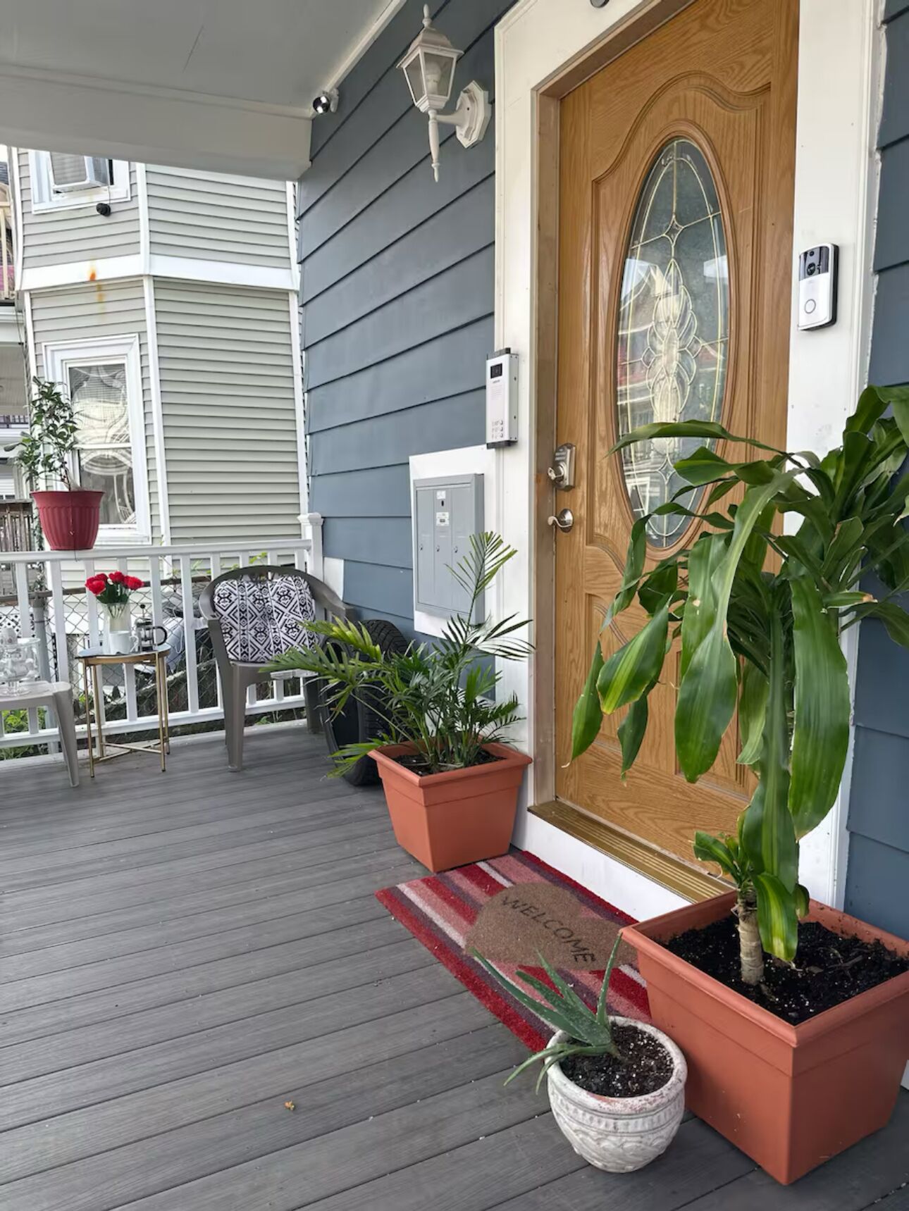 A wooden front door with a welcome mat, potted plants, and a porch with a table and chairs.