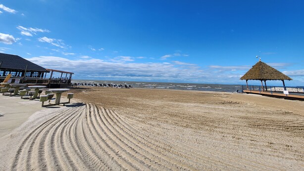 Ein Strand mit einem Strohdachschirm, Liegestühlen und einem Restaurant mit Außenbereich.