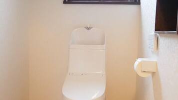 A small, white toilet in a corner with a wooden floor, a window above, and a wall-mounted electrical outlet to the right.