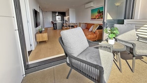 Una sala de estar moderna con un sofá, sillas y un comedor. Se observa una cocina al fondo y un balcón con mobiliario exterior.