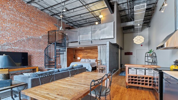 A loft-style room with a wooden dining table, a sofa, a TV, and a staircase leading to an upper level.