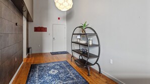 A hallway with wooden flooring, a blue patterned rug, a metal shelf with decorative items, and a hanging light fixture.