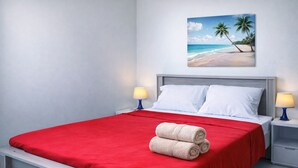 Un lit bien fait, recouvert d’une courtepointe rouge, avec deux oreillers blancs et des serviettes enroulées posées sur le côté. Un tableau représentant une scène de plage est accroché au mur, au-dessus du lit.