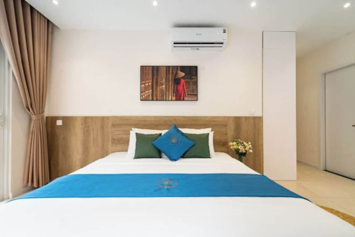 A hotel room with a bed, a wall-mounted air conditioner, a painting, and a window with curtains.