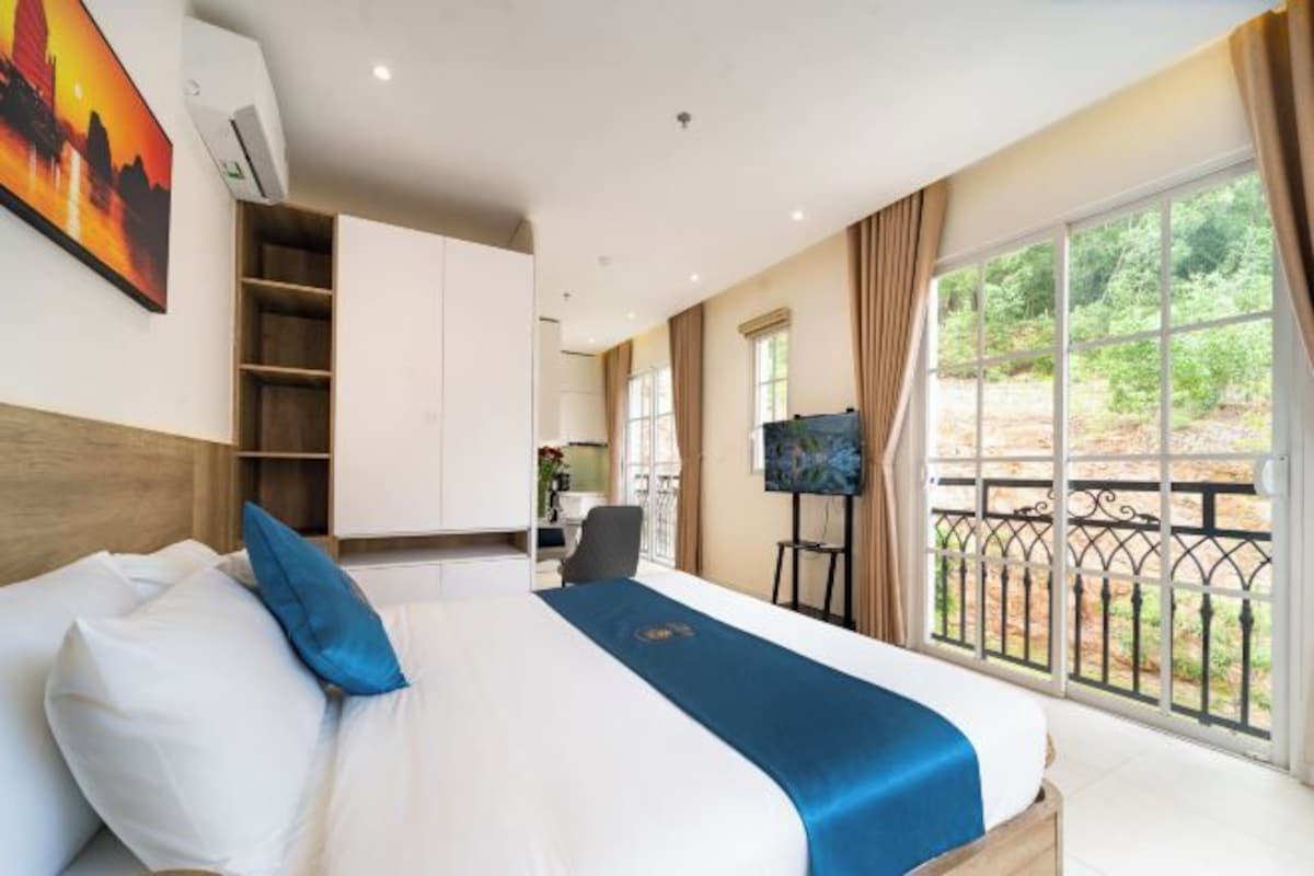 A hotel room with a large bed, a desk, and a balcony with a view of greenery.