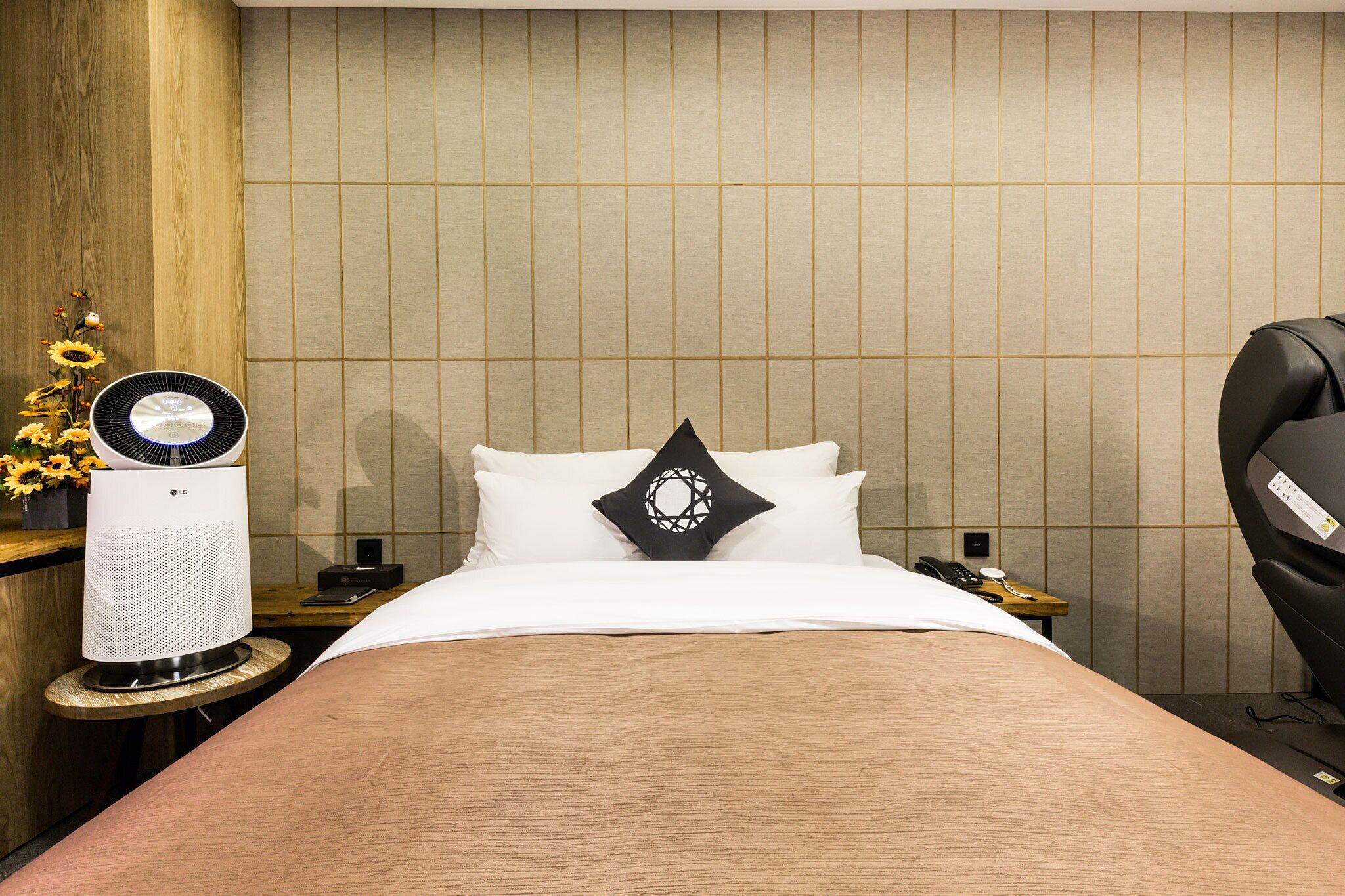 Habitación de hotel con cama, mesita de noche con teléfono y lámpara, calentador de pared y sillón de masaje.