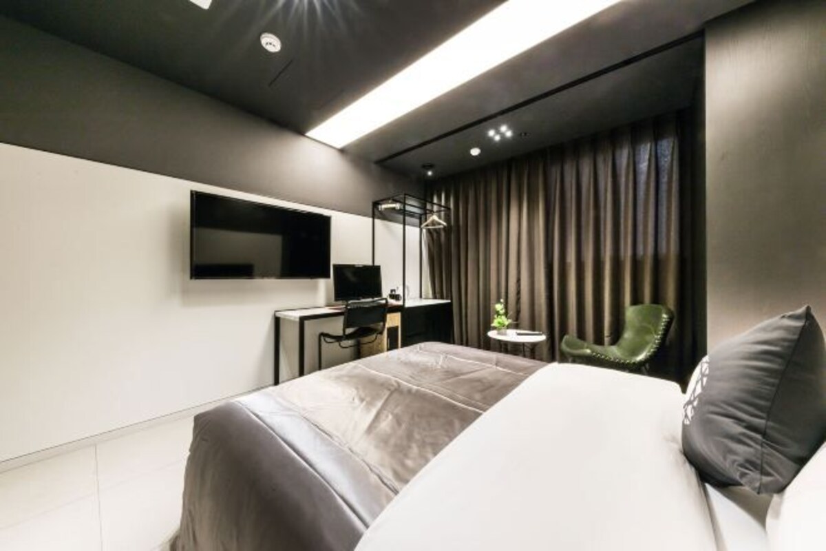 A modern hotel room with a large bed, a desk, a chair, a TV, and a small table.
