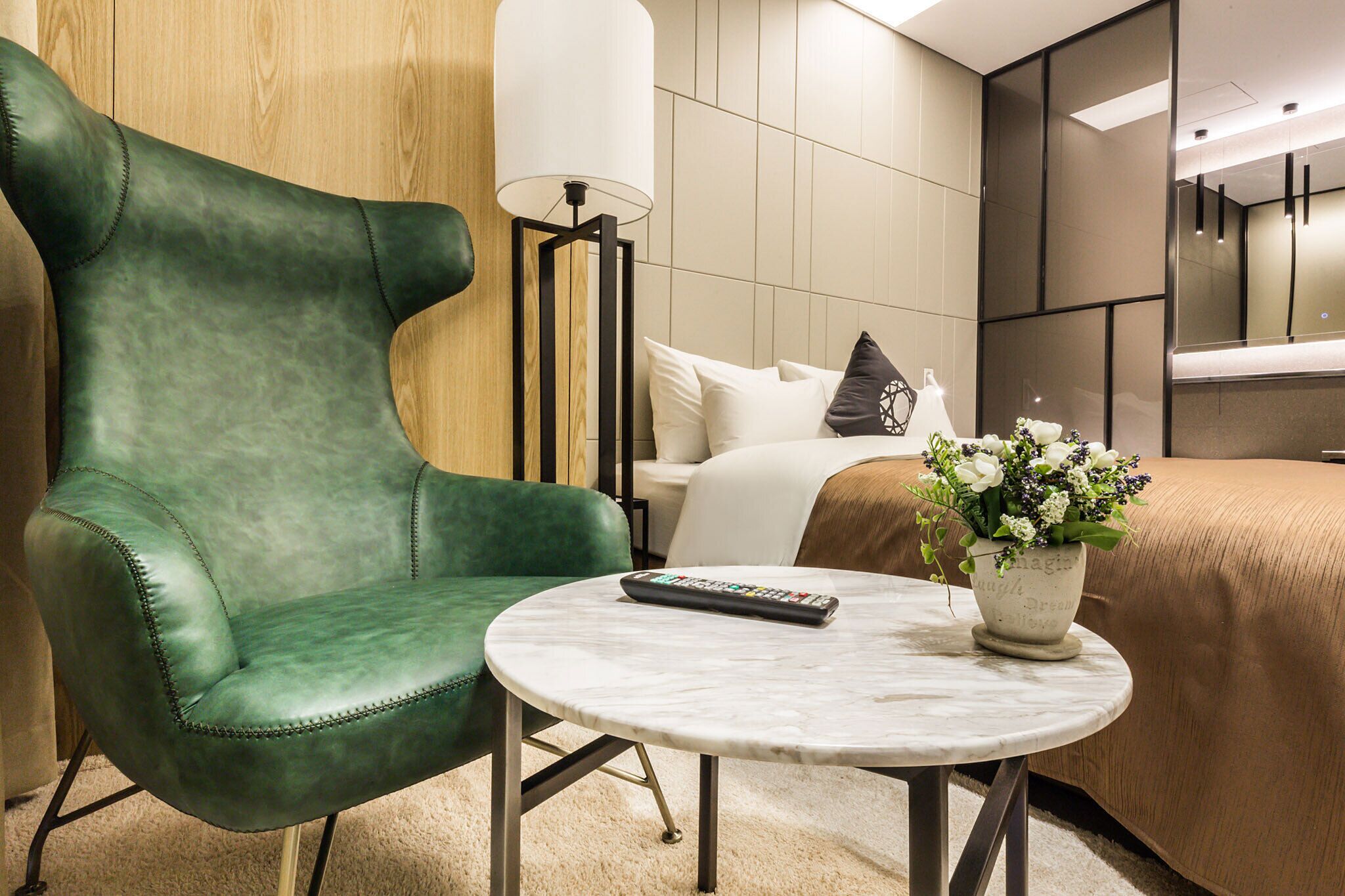 A hotel room with a green leather armchair, a round marble table with a remote control, a bed with a beige cover, and a potted plant with flowers.