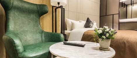 A hotel room with a green leather armchair, a round marble table with a remote control, a bed with a beige cover, and a potted plant with flowers.