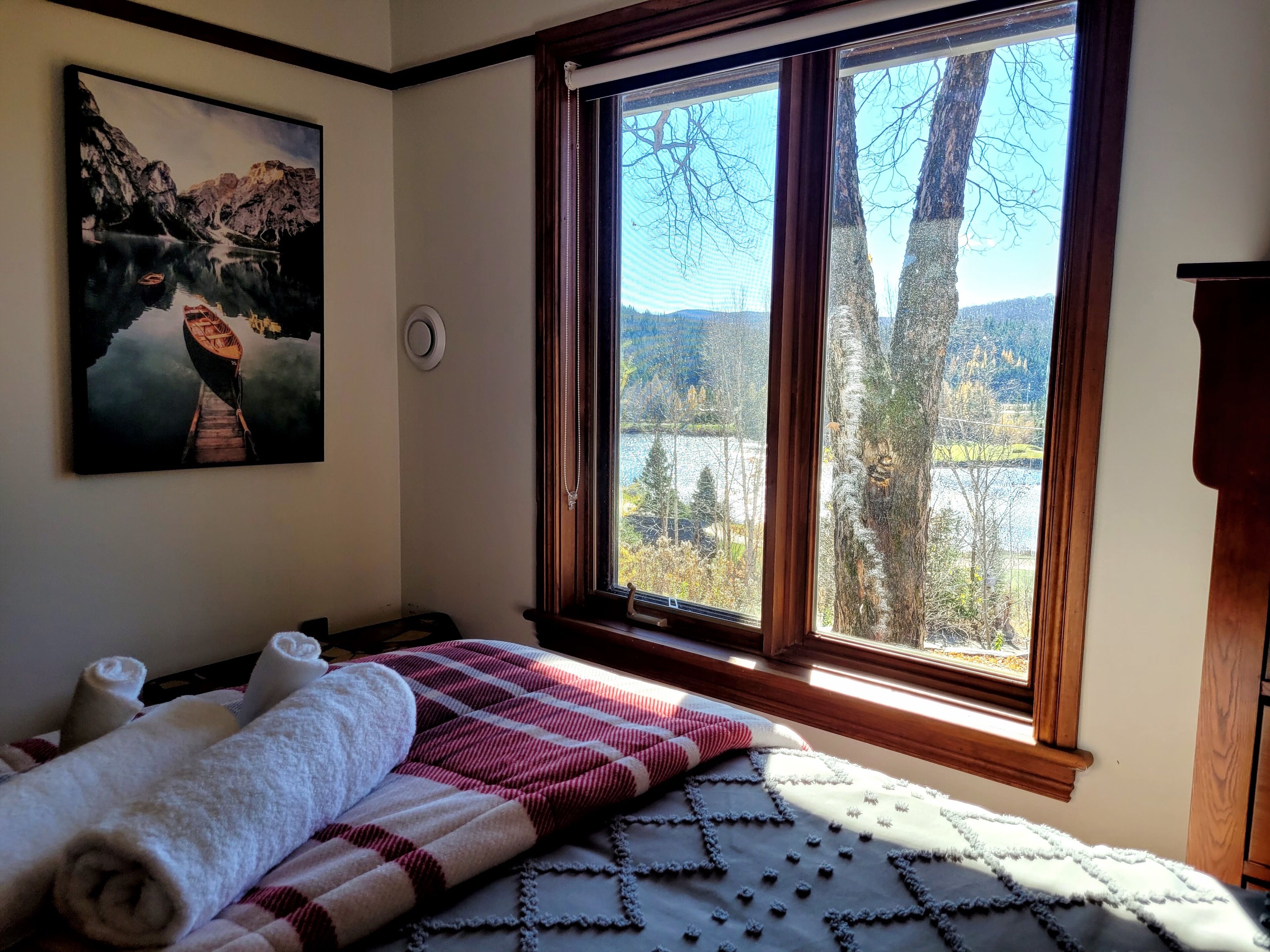A bedroom with a large window, a painting of a mountain lake, and a bed with towels and a quilt.