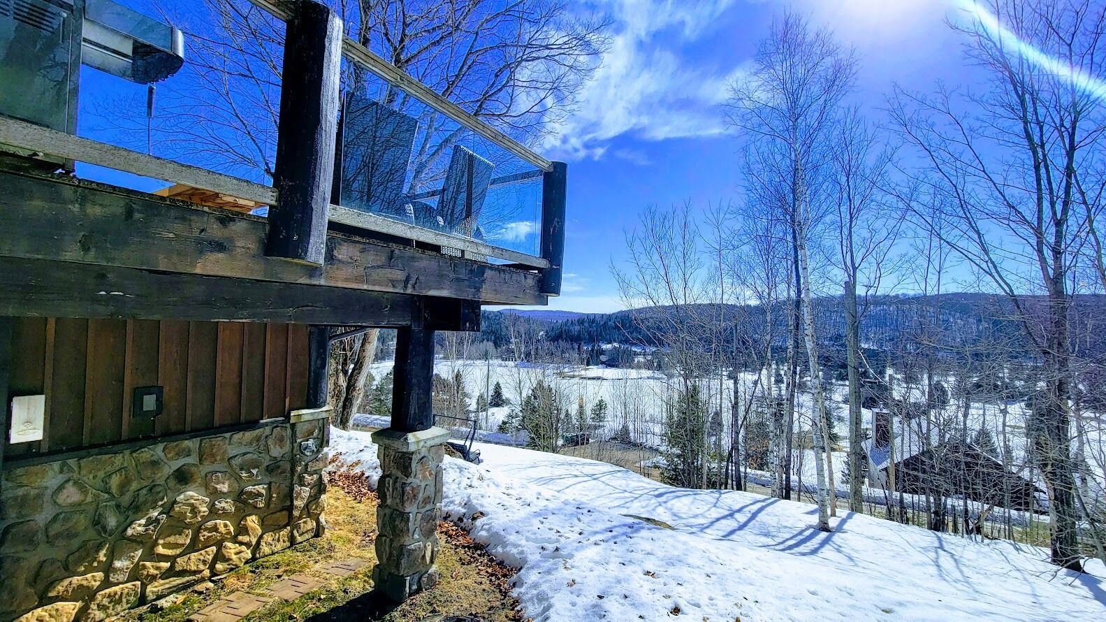Un bâtiment moderne doté d’un mur en pierre et d’une terrasse en bois, offrant une vue sur un paysage enneigé, avec des arbres et des maisons au loin.
