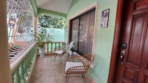 Un balcón con baranda verde, plantas en macetas, sillas de mimbre y una puerta de madera.