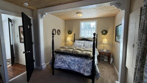 A bedroom with a four-poster bed, a window with a view, a small bedside table with a lamp, and a door leading to another room.