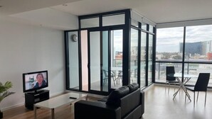 A modern living room with a black sofa, a small coffee table, and a TV on a stand. There is a large window with a view of the city. - RNR on Franklin-Triple Room with Balcony (Adelaide)