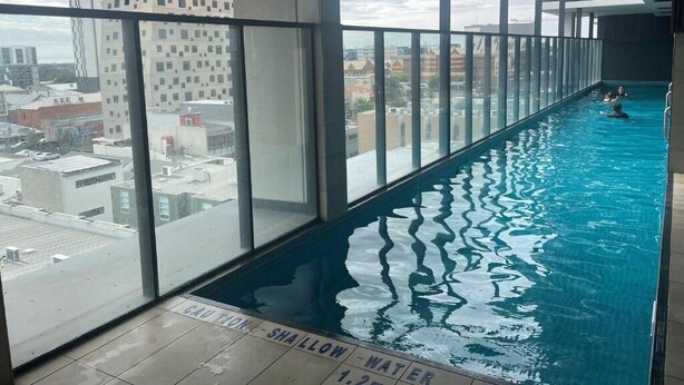 A long indoor swimming pool with clear water, surrounded by large windows offering city views. - RNR on Franklin-Triple Room with Balcony (Adelaide)