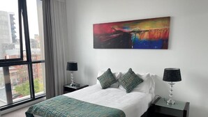 A hotel room with a large bed, two bedside tables with lamps, a colorful abstract painting on the wall, and a view of buildings through a large window. - RNR on Franklin-Triple Room with Balcony (Adelaide)