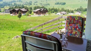 A terrace with a wooden railing, a table with a colorful chair, and a view of a green valley with buildings and mountains in the background. - Les Bluets by Interhome (Les Mosses)