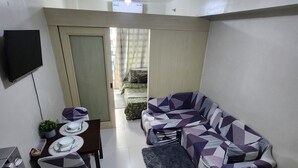A modern living room with a sofa, a dining table, and a TV mounted on the wall. - Cozy 1BR at Sea Residences Near MOA - Wifi & Netflix (Pasay)