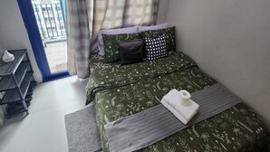 A bedroom with a bed, a small table, a shelf, and a view of a building through a window. - Cozy 1BR at Sea Residences Near MOA - Wifi & Netflix (Pasay)