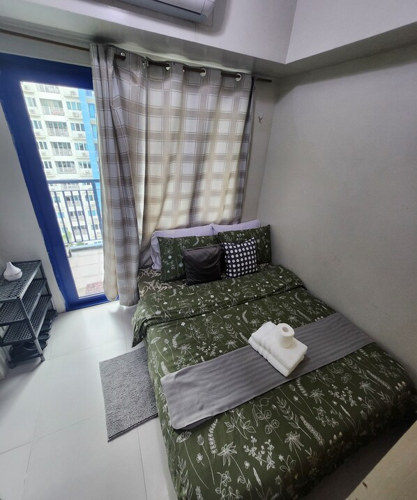 A bedroom with a bed, a small table, a shelf, and a view of a building through a window.