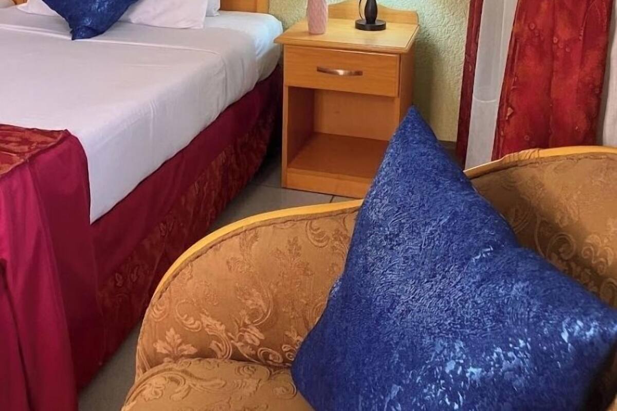 A hotel room with a bed, a nightstand with a lamp, a small potted plant, and two chairs.