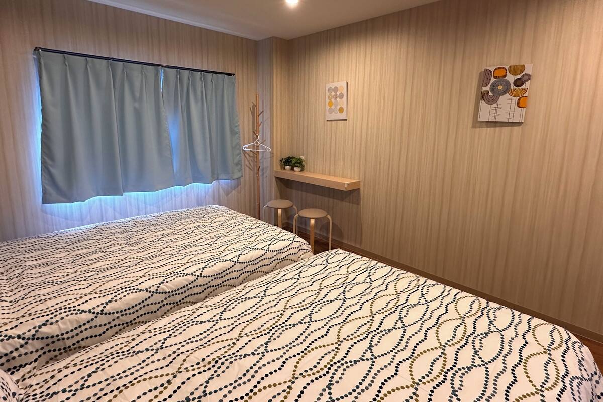 Un dormitorio con cama, ventana con cortinas, una pequeña estantería con objetos decorativos y un cuadro abstracto geométrico en la pared.