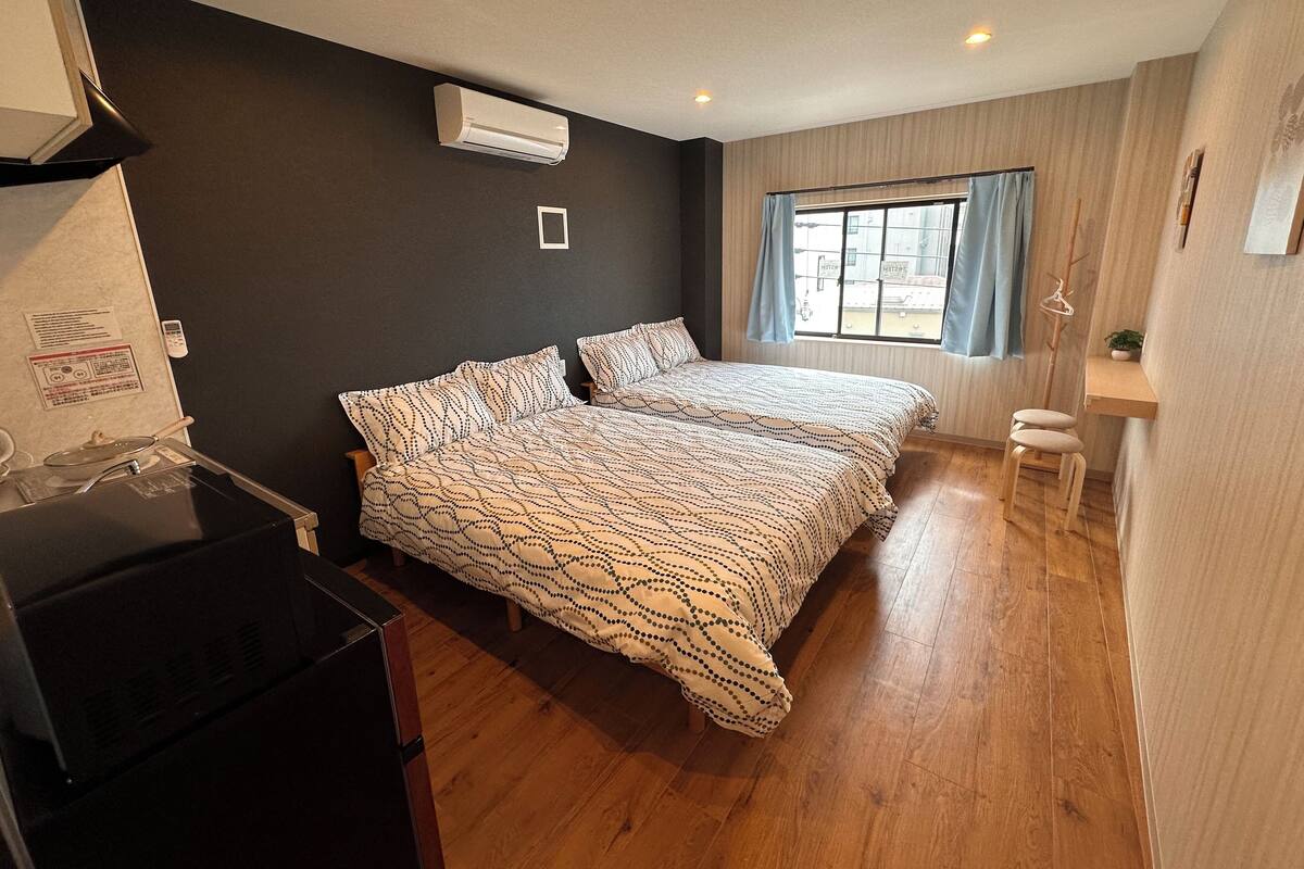 Un dormitorio con cama, una mesita, una silla, aire acondicionado y una ventana con cortinas.