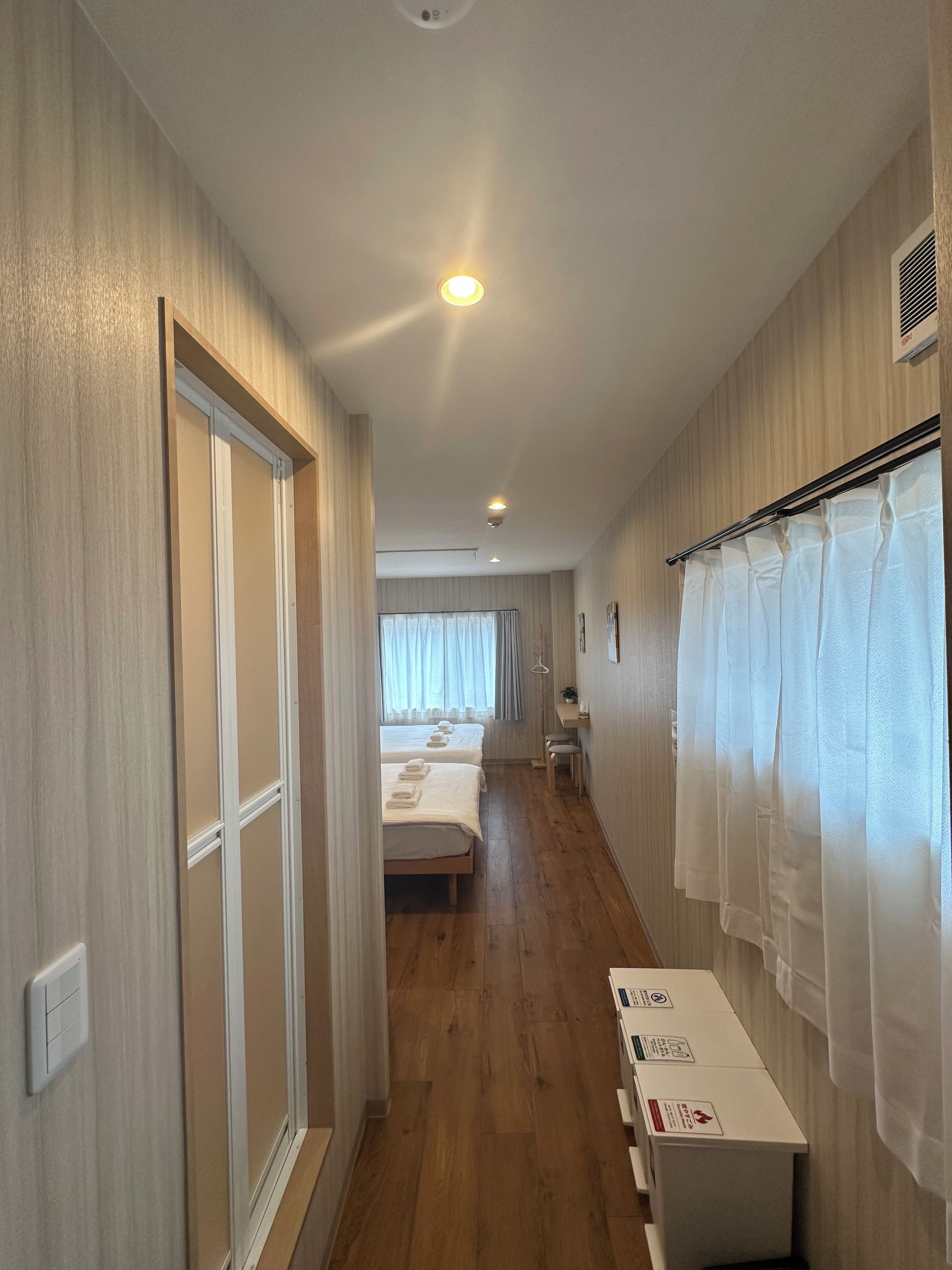 A hotel corridor with wooden flooring, a bed with white linens, a bedside table, and a window with curtains.