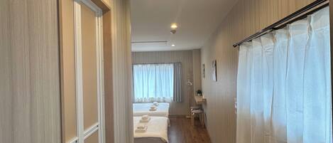 A hotel corridor with wooden flooring, a bed with white linens, a bedside table, and a window with curtains.
