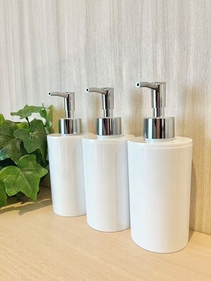 Tres dispensadores cilíndricos blancos con tapas metálicas, sobre una superficie de madera frente a una pared con textura.