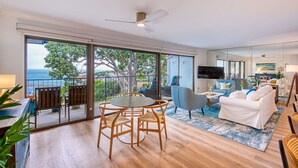 Uma sala de estar espaçosa com varanda vista para o mar, uma mesa de jantar com cadeiras, um sofá e uma televisão de tela plana fixada na parede.