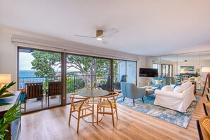 Un'ampia zona giorno con balcone vista mare, un tavolo da pranzo con sedie, un divano e una TV a schermo piatto fissata alla parete.