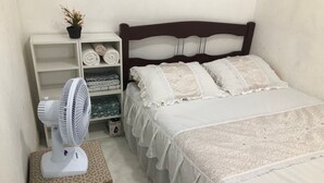 Un dormitori ben organitzat amb un llit, almoines, una taula de nit amb un ventilador i una estant amb tovalloles doblegades.