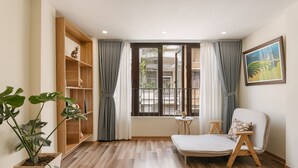 A room with a wooden floor, a white sofa, a small round table, a bookshelf, and a large window with curtains.