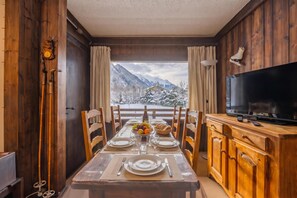A dining area with a table set for two, a television on a wooden cabinet, and a view of snow-covered mountains through the window.