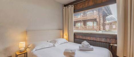 A hotel room with a bed, pillows, a nightstand with a lamp, and a window with curtains.