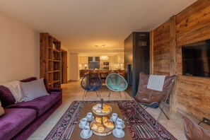 Een gezellige woonkamer met een paarse bank, een bijzettafeltje met gebak en kopjes, en een houten boekenkast.