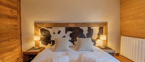 Un dormitorio con cabecera de madera, dos mesitas de noche con lámparas, cama con ropa de cama y toallas blancas, y un detalle decorativo con estampado de piel de vaca.