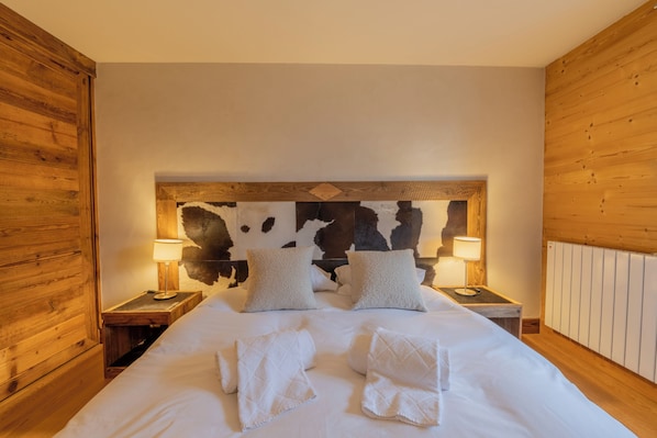 A bedroom with a wooden headboard, two bedside tables with lamps, a bed with white linens and towels, and a cowhide headboard accent.