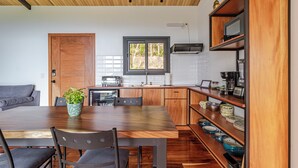 Een moderne keuken met houten kasten, een eettafel en stoelen. Er is een raam met uitzicht op groen.