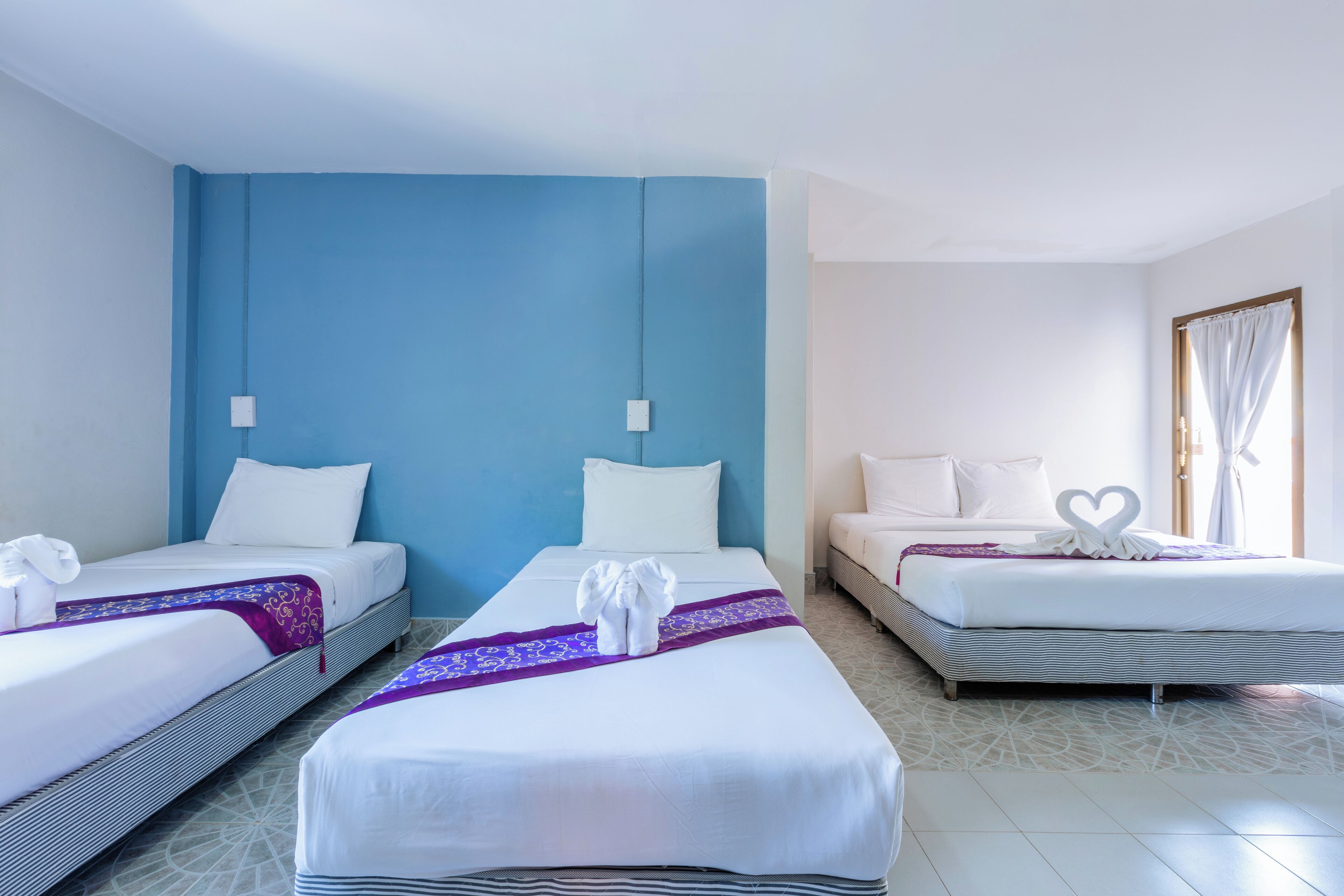 A hotel room with two beds, a blue accent wall, and a large window with curtains.