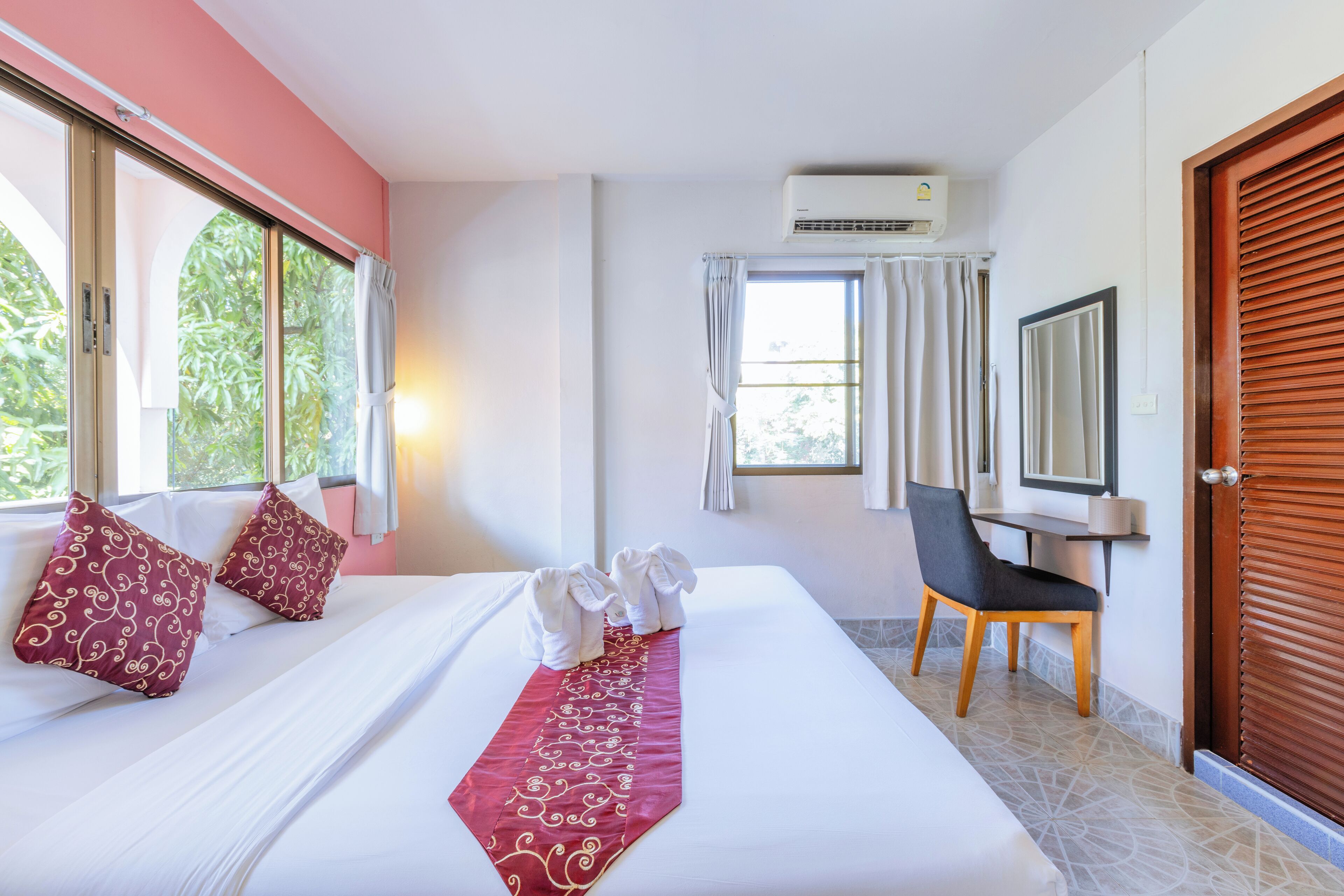 A hotel room with a bed, a desk, and a chair. There is an air conditioning unit on the wall, a window with curtains, and a door with a wooden frame.